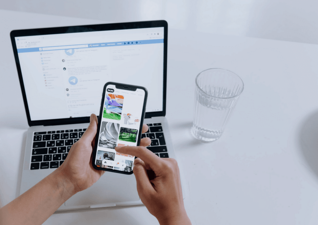 Person using Pinterest on cellphone with laptop on table showing Facebook page