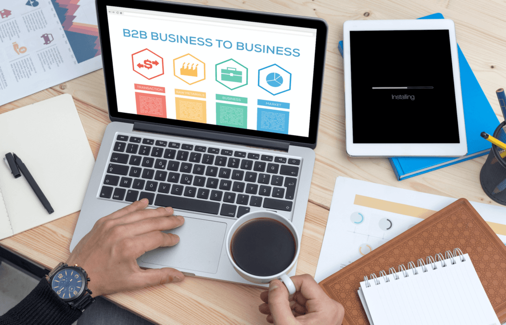 Person using laptop with B2B page at desk with tablet, documents, and coffee cup on table.