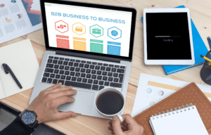 Person using laptop with B2B page at desk with tablet, documents, and coffee cup on table.