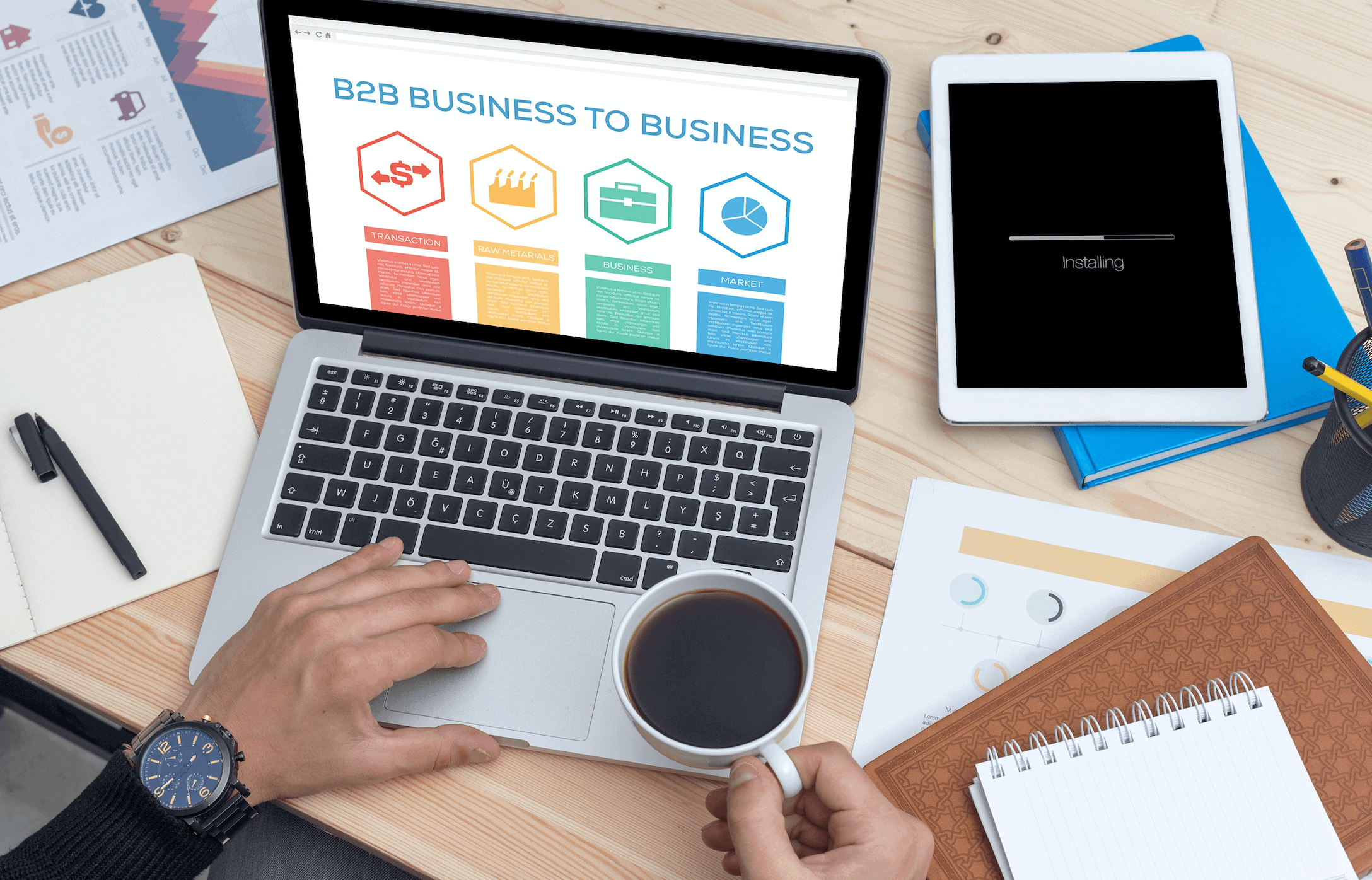 Person using laptop with B2B page at desk with tablet, documents, and coffee cup on table.